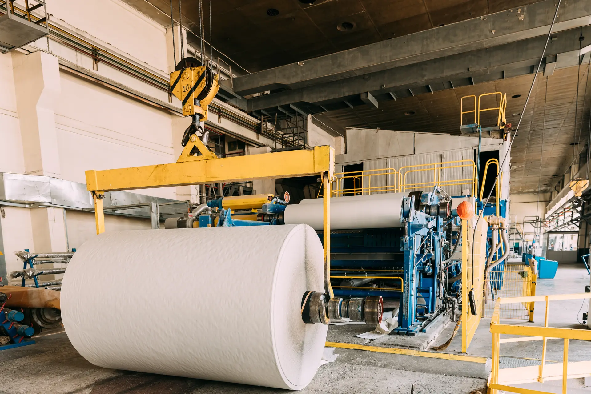 Große Papierrolle am Kran vor der Papiermaschine in einer Produktionshalle